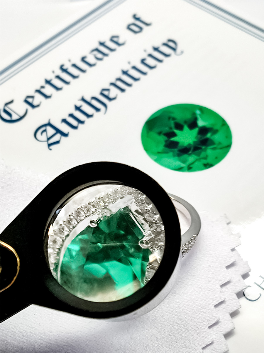Loop magnifying a Chatham lab-grown emerald set in a white gold ring with lab-grown diamonds and a Chatham Certificate of Authenticity behind it